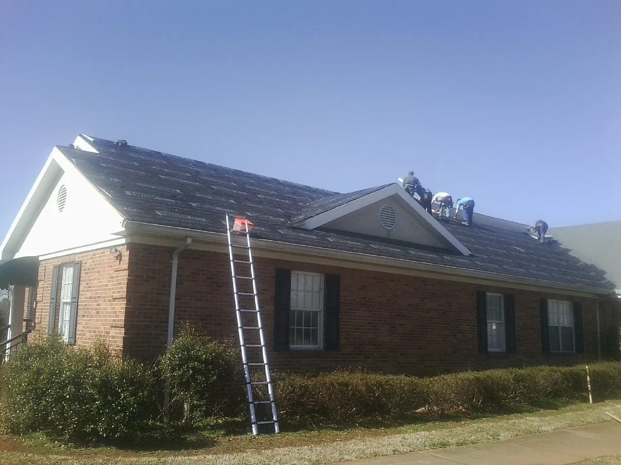 Crew tearing shingles off a brick ranch roof with a ladder set — Emergency Roof Repair work by our crew in West Olive