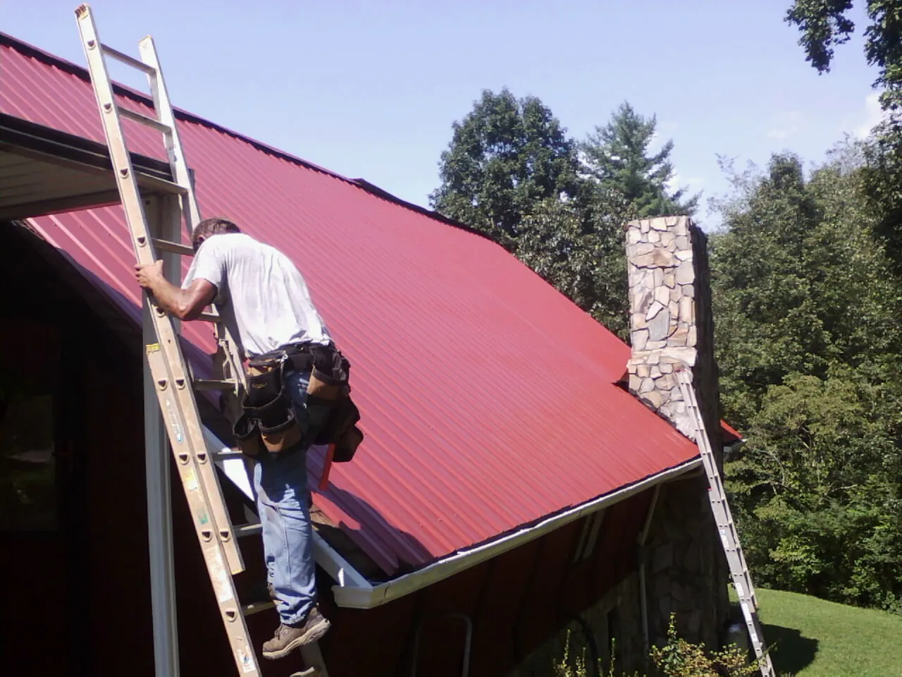 Roofer on a ladder beside a red metal roof and stone chimney — Soffit & Fascia Repair by Roofing Brothers in West Olive